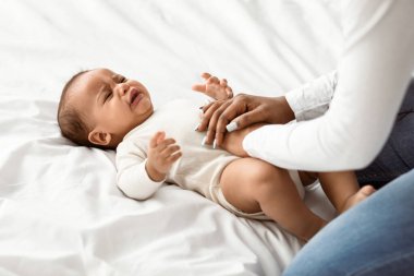 Tahrik olmuş Afrikalı Amerikalı küçük bir bebeğin portresi. Ağlayan ve sızlanan, kolik ya da mide ağrısı çeken, beyaz çarşafların üzerine uzanmış, yardım için karın masajı yapan genç bir anne.