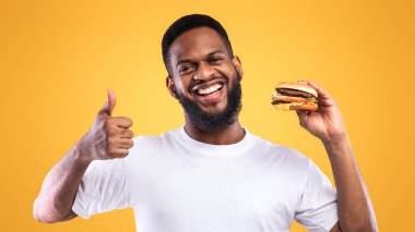 Abur cuburu severim. Elinde Burger tutan Afrikalı Adam, Sarı Stüdyo Arkaplanındaki Kameraya Gülümsüyor. Çizburgerleri Onaylayan Siyah Adam Fast food 'un tadına bakıyor.