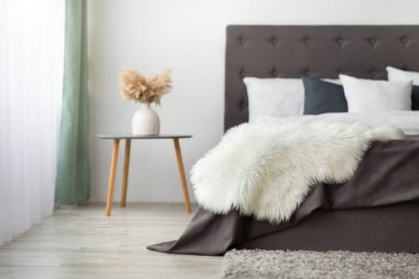 Boho ve İskandinav tarzında modern rahat şık yatak odası. Kahverengi battaniyeli yatak, yerde yumuşak halı vazodaki kuru bitkilerle masa ve gün ışığında gri duvar, düz yatak, boş alan.