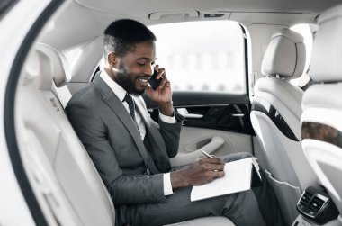 Afro işadamı arabada not defteri ile işlerini ayarlıyor, telefonda konuşuyor
