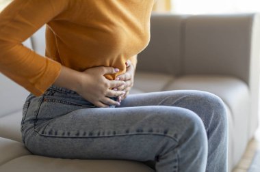 Tanımlanamayan bir kadın evde karın ağrısı çekiyor. Genç bir kadın oturma odasında koltukta otururken karnına dokunuyor. Rahatsızlık ve endişe gösteriyor.