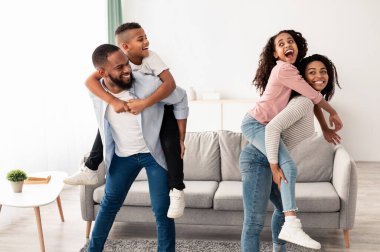 Pozitif siyah anne ve baba çocuklarıyla eğleniyor oturma odasında oynuyor, gülümseyen oğul ve kızı sırtında taşıyor. Dört kişilik Afro-Amerikan ailesi birlikte vakit geçirmekten zevk alıyor.