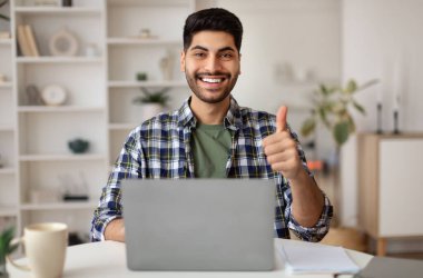 Hoşuma gitti. Mutlu Orta Doğulu Adam 'ın portresi, dizüstü bilgisayarla oturmuş işaret parmaklarını gösteriyor, el hareketlerini gösteriyor, çalışıyor ya da uzaktan çalışıyor, kameraya poz veriyor, seçici odaklanıyor.