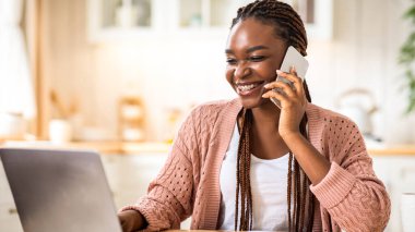 Uzaktan kumanda. Mutfakta cep telefonu ve dizüstü bilgisayar kullanan gülümseyen Afro-Amerikan Serbest Çalışan Kadın. Bilgisayarla çalışan ve cep telefonuyla konuşan mutlu siyah girişimci.