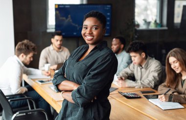 Başarılı bir insan. Kendinden emin gülümseyen Afrikalı Amerikalı iş kadınının portresi ofiste masaya yaslanmış, kollarını kavuşturmuş ve kameraya bakıyor, arka planda çalışan meslektaşları
