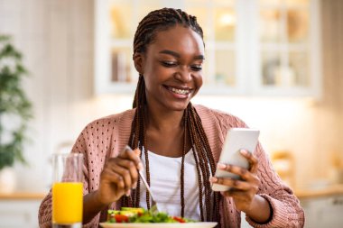 Mutfakta Kahvaltı Yaparken Akıllı Telefon Kullanan Genç Gülümseyen Siyah Kadın, Afro-Amerikan Kadın Cep telefonuyla sosyal ağları kontrol ediyor, İnternet 'ten alışveriş yapıyor ve Lezzetli Yemekler, Serbest Alan' ın tadını çıkarıyor.