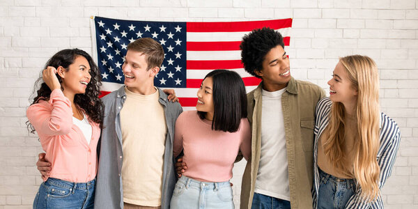 Acquaintance and first day in language courses or student exchange. Happy young international people look at each other, on white brick wall background with big USA flag, copy space, studio shot