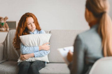 Depresyon Gençlik Konsepti. Ofiste ya da evde üzgün bir kızla çalışan kadın çocuk psikoloğu, aile sorunları hakkında konuşan, endişe duyan, kanepede oturan, yastığa sarılan bir genç.