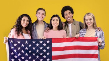Uluslararası bilgi programı, İngilizce öğrenme, değişim öğrencileri. Çok ırklı genç mutlu öğrenciler büyük ABD bayrağı tutuyorlar ve kameraya bakıyorlar, turuncu arka planda izole edilmiş, boş alan, stüdyo çekimi