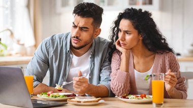 Endişeli Ortadoğu Eşleri, kahvaltıda laptopta Haberleri İzlerken Sinirli Arap Eşleri Sabah Yemeklerini yerken Bilgisayar ekranına bakıyor, Bilgiye Duygusal Tepkiler Veriyor