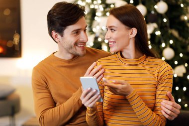 People And Technology Concept. Cheerful millennial couple sitting on couch at home using cell phone choosing what to buy together, excited guy embracing lady pointing at screen, sharing gadget