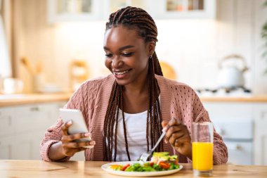 Gülümseyen siyah kadın evde kahvaltı yaparken akıllı telefon kullanıyor, neşeli Afro-Amerikan kadın masada oturuyor, cep telefonundan mesajlar okuyor ve yemek yiyor, bedava yer açıyor.