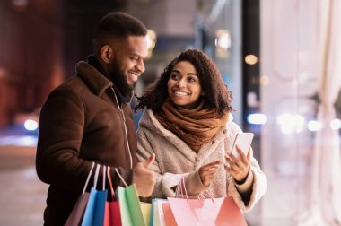 Mevcut Seçiliyor. Gülümseyen Afro-Amerikan kadın cep telefonunu erkeğe gösteriyor, cihazı işaret ediyor, alışveriş torbaları tutan adam, akşamları dışarıda duruyor. Teklif, Perakende ve Satın Al