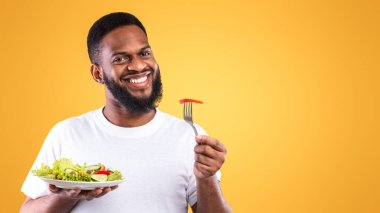 Afro-Amerikalı neşeli adam tabak ve çatalla salata yiyor. Sarı stüdyonun arkasında kameraya gülümsüyor. Sağlıklı Vejetaryen Yemeği, Erkek Beslenme ve Kilo Verme Konsepti