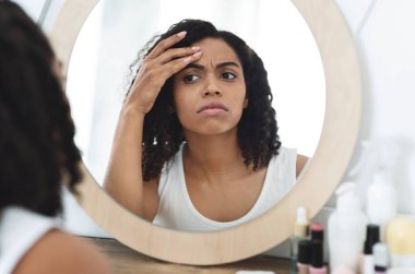 İnce çizgiler ve kırışıklıklar. Endişeli siyahi kadın aynaya bakıyor. Yüzündeki yaşlanma işaretlerini kontrol ediyor, seçici odaklanıyor.