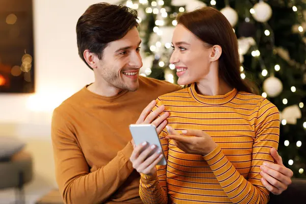 People And Technology Concept. Cheerful millennial couple sitting on couch at home using cell phone choosing what to buy together, excited guy embracing lady pointing at screen, sharing gadget