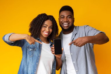 Yeni mobil uygulama. Neşeli Afro-Amerikan çifti ellerinde tuttukları ve siyah ekranlı akıllı telefonları göstererek sarı arka planda birlikte poz veriyorlar.
