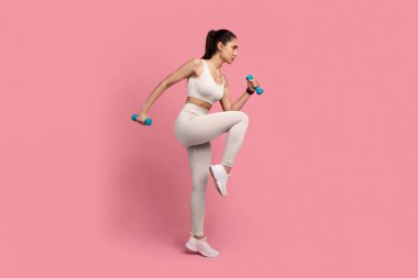 Eğitim Konsepti. Spor Giyim 'de iki dambılla spor yapan ve Pembe Stüdyo arka planında izole edilmiş Bacak kaldıran Kendine Güvenen Genç Bayan' ın tam uzunlukta portresi. Enerji ve Spor