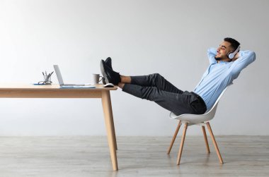 Dinlen ve rahatla kavramı. Sandalyeye oturup müzik, sesli kitap, kablosuz kulaklıkla podcast, masaya yaslanmış ayaklar masada ve eller başının arkasında oturarak mutlu bir Arap.