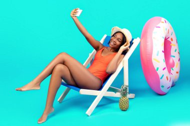 Muhteşem siyahi kadın, mavi arka planda şezlongda akıllı telefonuyla selfie çekiyor. Boş yeri olan afiş. Pozitif genç bayan bikinisiyle fotoğraf çekiyor, serin yaz tatili geçiriyor.