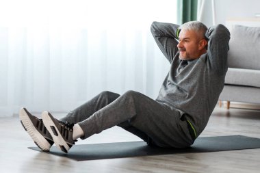 Gri spor giyim eğitiminde olgun bir adam, spor kıyafetlerini giyip, spor paspasına uzanıp mekik çekiyor, karın kasları üzerinde çalışıyor, tripodda modern akıllı telefonu kullanıyor, online fitness dersleri izliyor, bedava uzay dersleri alıyor.