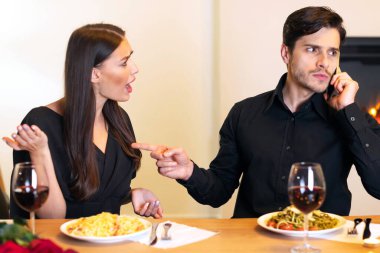 Relationship Conflicts Concept. Annoyed angry woman looking and yelling at her man who talking on cellphone and showing her finger to shut up and to wait while having date in restaurant