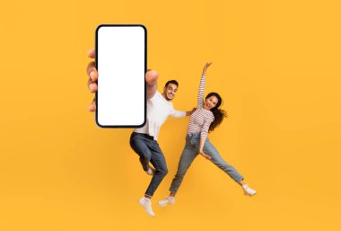 Sarı Stüdyo Arkaplanı Üzerinde Büyük Boş Akıllı Telefonla Atlayan Neşeli Genç Arap Çift, Mobil Tasarım, Mobil Tasarım İçin Boş Cep Telefonu Gösteren Mutlu Ortadoğulu Erkek ve Kadın