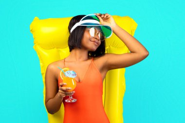 Sarı şişme lilonun üzerine uzanmış, mavi stüdyo arka planında meyve kokteyli içen mayolu, uzun boylu, rahat bir zenci kadın. Seksi Afro-Amerikan kadın yaz nezlesi geçiriyor.