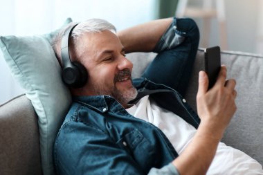 Modern kablosuz kulaklıklı neşeli, gri saçlı, orta yaşlı adam akıllı telefondan müzik dinliyor, fotokopi odası, yakın çekim. Kulaklıkla cep telefonundan video izleyen mutlu olgun bir adam.