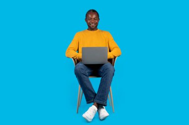 Sarı kazaklı mutlu Afrikalı orta yaşlı adam, mavi stüdyo arka planında modern laptop kullanıyor, klavyeye yazıp gülümsüyor, çalışıyor ya da oyun oynuyor, boşluğu kopyalıyor.
