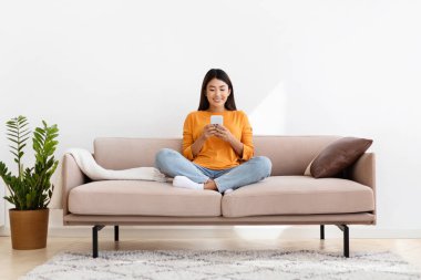 Milenyum Asyalı gülümseyen bayan akıllı telefon tutuyor, rahat oturma odasında, ev içinde kanepede oturuyor. Filipinli kadın çevrimiçi kitap veya blog okuyor cep telefonu, kopyalama alanı, tam uzunluk