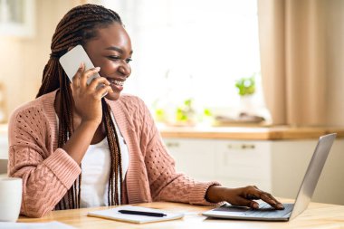 Telekomünikasyon konsepti. Siyah kadın cep telefonu kullanıyor ve mutfakta dizüstü bilgisayarla çalışıyor. Gülümseyen Afrikalı Amerikalı Serbest Çalışan Kadın Cep telefonuyla konuşuyor ve uzaktan kumandalı bilgisayarla klavye çalıyor.