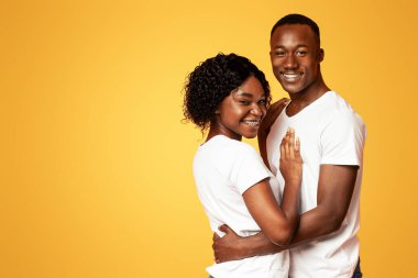 Beyaz tişörtlü, turuncu stüdyo arka planında kameraya sarılıp gülümseyen sevimli Afro-Amerikan genç çifti mesaj ya da reklam için fotokopi alanının yanında duruyorlar.