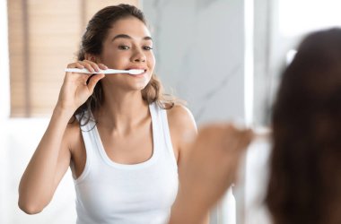 Neşeli kadın diş fırçası ve diş macunuyla diş fırçası fırçalıyor aynanın yanında duruyor ve modern banyoda diş bakımı rutini yapıyor. Diş Sağlığı Ürünleri Reklamı. Ağız Temizliği Konsepti