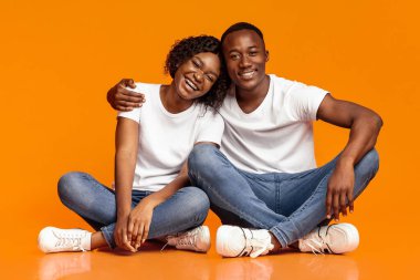 Kaygısız Afrikalı Amerikalı çift yerde oturuyor ve sarılıyorlar, kameraya bakıyorlar ve gülümsüyorlar, boşluk. Genç mutlu siyah adam ve kadın turuncu stüdyo arka planında birlikte poz veriyorlar.