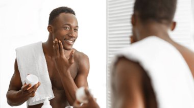 Mutlu Siyahi Adam Yüzü Nemlendirici Kullanıyor Kozmetik Krem Kavanozu, Yüzü İçin Bakıyor Evdeki Modern Banyo 'da Üstsüz Ayna Yapıyor. Erkek derisi ve güzellik rutini. Panorama