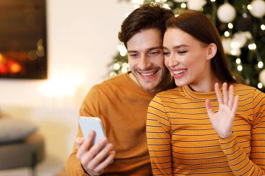 Remote Communication. Portrait Of Smiling Couple Holding And Using Mobile Phone Sitting On Sofa In Living Room, Waving Hands To Screen Webcam. Two Happy People Making Video Call With Family Or Friends