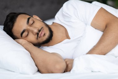 Orta Doğulu yakışıklı bir gencin evde rahat bir yatakta uyuduğu, rahat bir yatak odasında rahat bir şekilde uyuyan, elleri başının altında yumuşak yastığın üzerinde uzanan bir Arap 'ın yakın plan fotoğrafı.