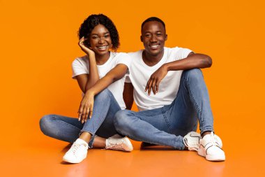 Stüdyoda poz veren, turuncu arka planda, fotokopi alanında birlikte oturan sevimli siyah çift. Gülümseyen Afrikalı Amerikalı genç kadın mutlu erkek arkadaşıyla bağ kuruyor, birlikte vakit geçiriyor.