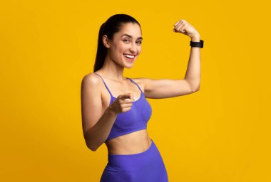 Başarabilirsin. Spor kıyafetli heyecanlı genç bir kadının portresi, bisepsleri gösteriyor ve kamerayı işaret ediyor, turuncu sarı stüdyo arka planında izole edilmiş. Spor ve Sağlıklı Yaşam Tarzı