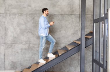 Bu resim, modern bir evde merdivenlerden çıkan bir adamı gösteriyor. Adam tesadüfen mavi gömlek ve kot pantolon giymiş ve elinde bir telefon tutuyor. Merdivenler tahtadan yapılmıştır.