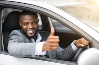 Takım elbiseli Afro-Amerikan iş adamının pahalı bir araba sürüşü, mutlu bir şekilde gülümsemesi ve başparmağını kaldırışı, yakın çekim. Genç siyahi girişimci başarılı bir iş toplantısından sonra arabayla evine dönüyor.