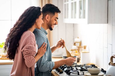 Birlikte Yemek Pişirme Konsepti. Genç Arap eşler mutfakta yemek hazırlıyor. Orta doğulu yakışıklı bir adam spatulayla yemek pişiriyor. Karısı da onu arkadan, yandan, serbest mekandan kucaklıyor.