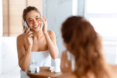 Her zaman temastayız. Banyoda beyaz havluya sarılı bir şekilde göz altı bandı takan mutlu kadın telefonda konuşuyor. Modern Kadın Yaşam Tarzı, Güzellik Rutini. Seçici Odaklanma