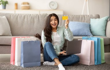 Aşırı neşeli Hintli kadın online mağazada büyük satışlardan dolayı mutlu hissediyor, dizüstü bilgisayar kullanıyor, kredi kartını gösteriyor, evde hediye çantalarıyla yerde oturuyor. Heyecanlı bayan içeride internetten mal alıyor.