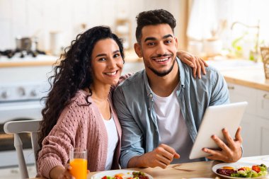 Mutlu Arap eşlerin portresi, kahvaltıda dijital tabletlere bakınıyor, Orta Doğu 'lu neşeli eşler ve modern Gadget mutfaktaki masada oturuyor ve lezzetli yiyeceklerin tadını çıkarıyor.