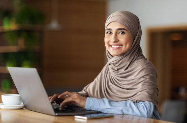 Kafe 'de çalışan tesettürlü neşeli Müslüman kadın dizüstü bilgisayar kullanıyor, kahve veya çay içiyor, fotokopi çekiyor. Yan bakışta gülümseyen Arap kadın dizüstü bilgisayarda yazı yazıyor, uzak işinden zevk alıyor.