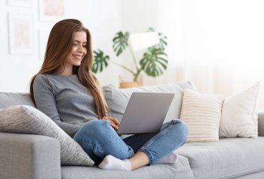 Evdeki kanepede dizüstü bilgisayar kullanan mutlu genç bayan arkadaşlarıyla sohbet ediyor, boşluğu kopyalıyor.