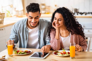 Genç Orta Doğulu çift, birlikte kahvaltı ederken dijital tabletten alışveriş yapıyor, gülümseyen genç Arap eşler mutfakta modern Gadget kullanıyor ve sağlıklı yiyeceklerin tadını çıkarıyor, yakın plan
