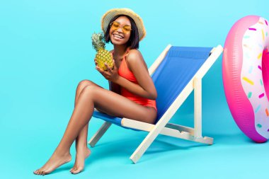 Güzel bir yaz tatili. Bikinili neşeli genç bir kadın ananas tutuyor, şezlongda oturuyor ve mavi stüdyo arka planında kameraya gülümsüyor. Siyah kadın yaz tatilinin tadını çıkarıyor.